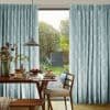Floor-length pleated pale-blue patterned curtains pulled aside, framing a glass patio door and softly filtering daylight; a wooden dining table set for breakfast, pendant lamp, chairs, and sunny garden view.