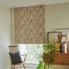 Roman fabric Roman shade with brown-and-cream leaf pattern, lowered and softly filtering daylight. Context: cozy living-room nook with wooden chair, cushion, sideboard, plants, and framed art.
L'ATELIER BOTANIQUE