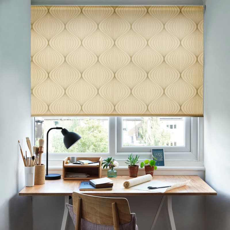 Patterned roller blind covering the upper two-thirds of a window, pale gold with curving geometric motifs — lowered to filter daylight — small wooden desk, lamp, art supplies and potted plants beneath.