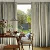 Floor-to-ceiling pleated striped curtains (gray, white, yellow) frame a central glass door, parted to reveal sunlight and a leafy backyard; foreground dining table set with pancakes, pendant lamp, wooden chairs.
