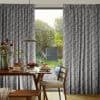 Floor-length pleated grey floral-patterned curtains, drawn apart at center, framing a sunlit garden; they filter soft light onto a wooden dining table set for breakfast with pendant lamp and chairs.
