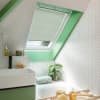 Pleated white cellular blind on an angled skylight, partially lowered and filtering daylight; sits above a green-framed attic bathroom with herringbone white tiles, bathtub, towel, potted plant, and toiletries.