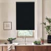Black roller blind covers the upper portion of a kitchen window, partially blocking daylight above a stainless sink, set against a countertop with vegetables, potted plants and framed wall art.