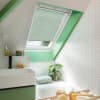 Mint-green pleated skylight blind, lowered and filtering daylight with a narrow open gap, in an angled bathroom window; white herringbone tiles, green trim, sink, tub, and potted plant nearby.