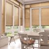 Tan horizontal Venetian blinds cover floor-to-ceiling windows and doors, lowered and filtering warm sunlight; bright dining sunroom with white wooden table and chairs, floral cushions, tiled floor and garden view.