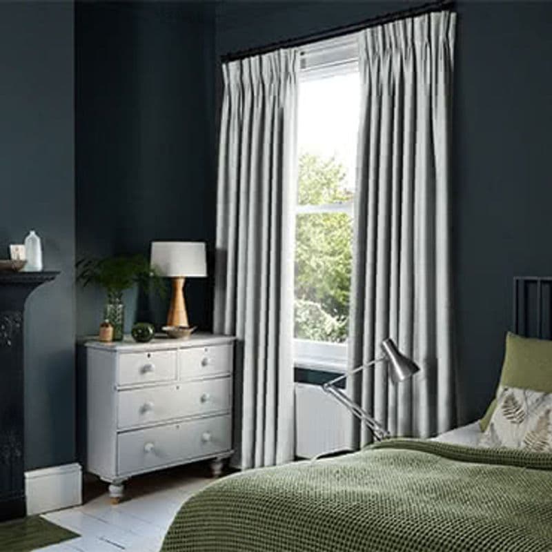 Pleated grey floor-length curtains on a black rod, pulled to each side and framing a tall sash window, admitting bright daylight; bedroom with dark teal walls, white dresser, green bedding.
