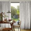 Pleated, patterned curtains with delicate leaf print, pulled slightly aside; filtering soft daylight through a glass door into a dining nook with wooden table, chairs, breakfast setting and garden view.
