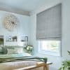 Grey textured Roman blind covering a window, lowered to block and filter light; in a brightly lit bedroom with green-accent bedding, built-in headboard niches, plants, and a feather wall hanging.