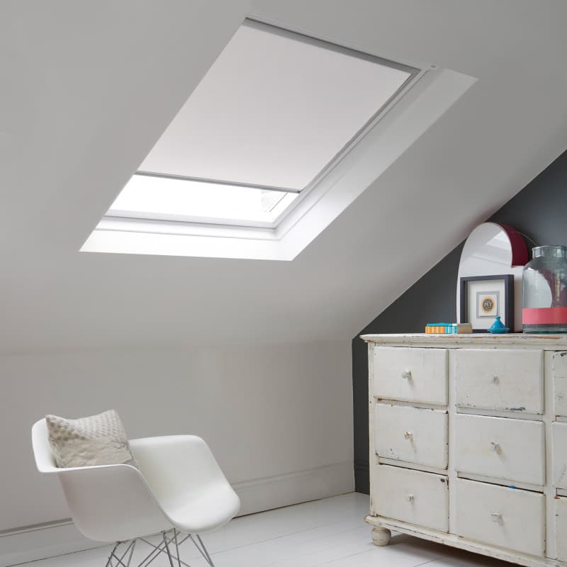 Room with white eaves wall, dark blue-grey wall to right with a shabby chic white drawer unit, a contemporary white chair, white painted floorboards and a skylight blind in lily white.