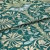 Teal-green woven fabric with pale blue and cream floral vines; folded across the frame, displaying textured weave and layered petals; close-up view under soft diffuse light on a flat surface.