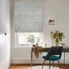 Floral-patterned Roman blind lowered over a sash window, gently filtering daylight; above a wooden desk with mirror, teal chair and vase of flowers in a bright home office.