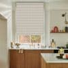 Beige textured roller blind, lowered across the window with horizontal weave; filtering soft daylight. Kitchen sink with brass faucet below, marble countertop, wooden cabinets, shelf with objects, brick courtyard outside.