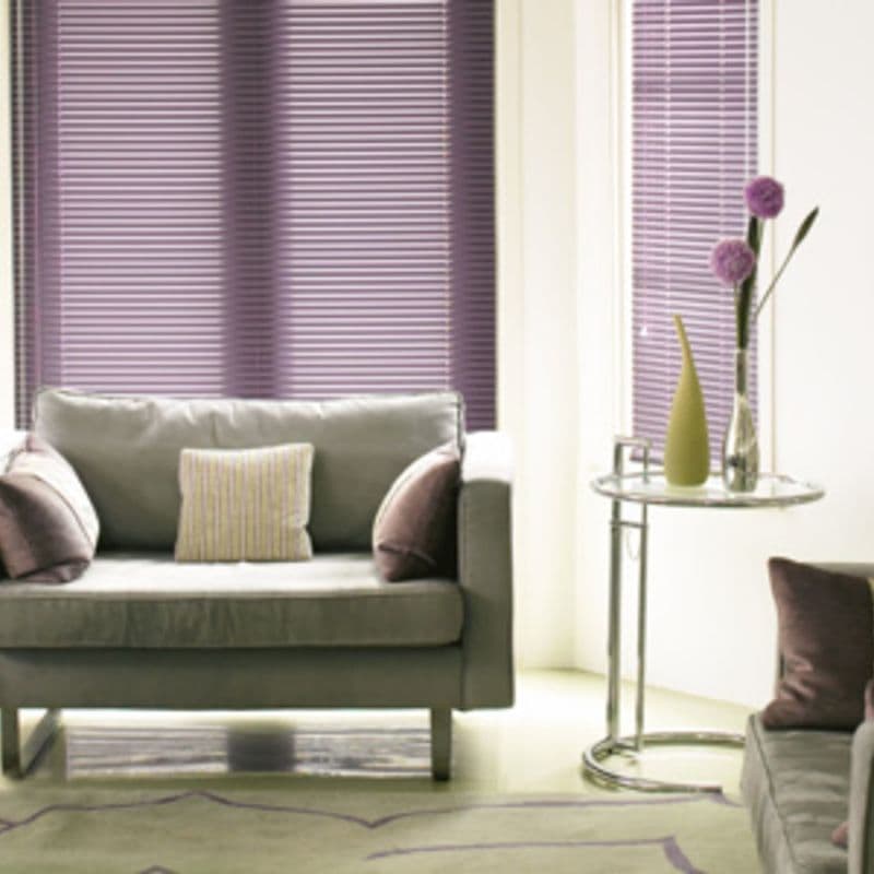 Purple horizontal blinds covering tall windows, fully closed and filtering soft light; a gray upholstered sofa with pillows faces a glass side table holding vases, in a lit living room.