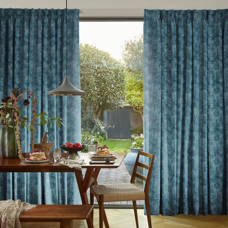 Teal textured full-length pleated curtains, parted slightly at center, framing a sliding glass door; filtering soft daylight into a dining nook with wooden table, chairs, pendant lamp and garden view.