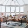 White pleated cellular blinds covering conservatory roof and tall bay windows, fully lowered and softly filtering daylight; a sunlit sitting room with beige sofa, chunky pink throw, and wooden coffee table.