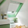 White pleated skylight blind, mounted inside a roof window frame, pulled down about two-thirds, filtering daylight; set above a green-accented bathroom with white herringbone tiles, sink, tub, plant, toiletries.