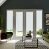 White roller blinds cover tall French doors, filtering light into a living room with a glass coffee table, chevron rug, green velvet armchairs, and glimpses of a sunlit garden outside.