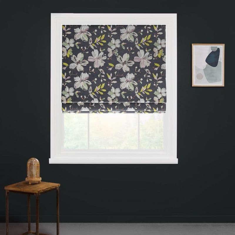 A floral Roman blind, dark background with blossoms and green leaves, is lowered across a white window, softly filtering daylight in dark room with side table and abstract wall art.