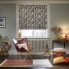 Patterned Roman blind (black leafy print) lowered over a white window, filtering daylight; in a cozy living room with leather butterfly chair, radiator, lamp, cushions, and wood floor.