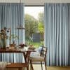 Striped, pinch-pleat floor-length curtains (blue, gray, white) parted at center, framing and softening light; a dining table with pendant lamp, chairs, and breakfast faces a sunlit backyard garden.