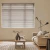 Horizontal Venetian blinds covering a large rectangular window, closed and filtering soft daylight through thin slats; living room with beige sofa, wooden coffee table, lamp, sculptural vase and open book.