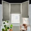 Grey fabric Roman blinds covering a three-panel bay window, partially lowered and filtering bright daylight into a white room with a radiator below, a wooden armchair, and a potted Monstera.