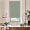 A muted green roller blind covers a window above a stainless faucet, with a marble countertop, wooden cabinets, salad bowl and potted plants flanking the sink in a bright kitchen.