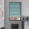 Teal zebra roller blind, lowered mid-window, with alternating opaque and sheer horizontal bands filtering daylight; set in a gray kitchen room with radiator beneath, countertop espresso machine and cactus prints.