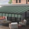 Green-and-light-green striped retractable awning, fully extended with matching valance; shading a wooden-deck patio with wicker chairs, round coffee table, and sliding glass doors beneath a brick upper wall.