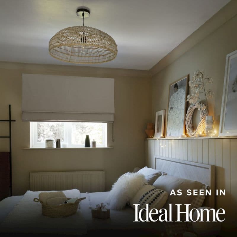 A white Roman blind lowered across a small rectangular window, diffusing soft daylight; in a neutral-toned bedroom with a woven pendant lamp, wooden headboard, cushions and a fairy-lit shelf with framed art and ornaments.
AS SEEN IN Ideal Home