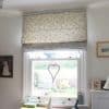 Cream fabric Roman blind with grey vine embroidery, lowered halfway and softly filtering daylight; hangs over a white-framed sash window in a bright room with photos, desk and garden view.
