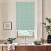 Patterned teal roller blind with small white daisies covers a kitchen window, filtering light above a sink and faucet; countertop holds bowls of greens, potted plants and a framed sketch.