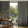 Floor-length pleated gray curtains, hung across a sliding glass door, parted at center; filtering soft daylight and framing a dining table set for breakfast and a leafy backyard view.