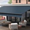 Blue striped retractable awning with matching valance extended over patio glass doors, shading a furnished terrace—wooden coffee table, cushioned chairs and bench—attached to a brick-faced modern house under daylight.