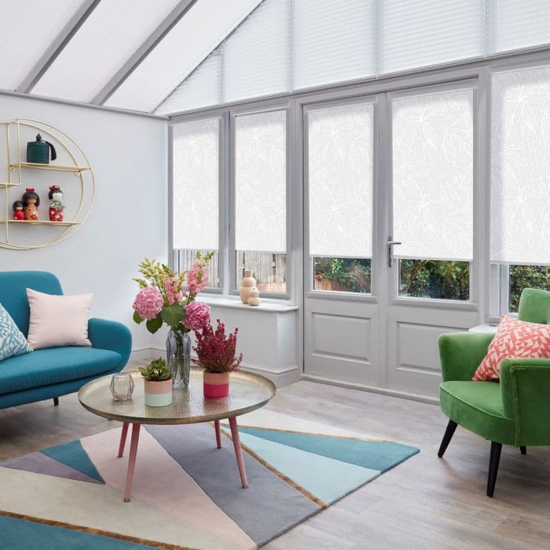 White patterned roller blinds and upper pleated blinds cover conservatory windows and French doors, lowered and filtering daylight; living room with teal sofa, green armchair, round coffee table and plants.