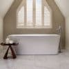 White plantation shutters cover a pitched-arched window, slats tilted to filter daylight; a freestanding white tub sits beneath with chrome floor-mounted faucet, marble floor, neutral sloped attic bathroom.