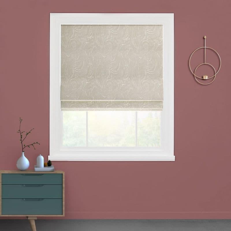Beige patterned Roman shade, lowered with visible bottom fold, filtering soft daylight; in a white-trim window of a mauve-walled room with a teal dresser and circular gold wall decor.
