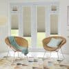 Beige pleated blinds fitted across French doors, sidelights and transoms, partially lowered to filter daylight with lower panels raised. Bright sitting room with woven chairs, cushions, patterned rug, lawn view.
