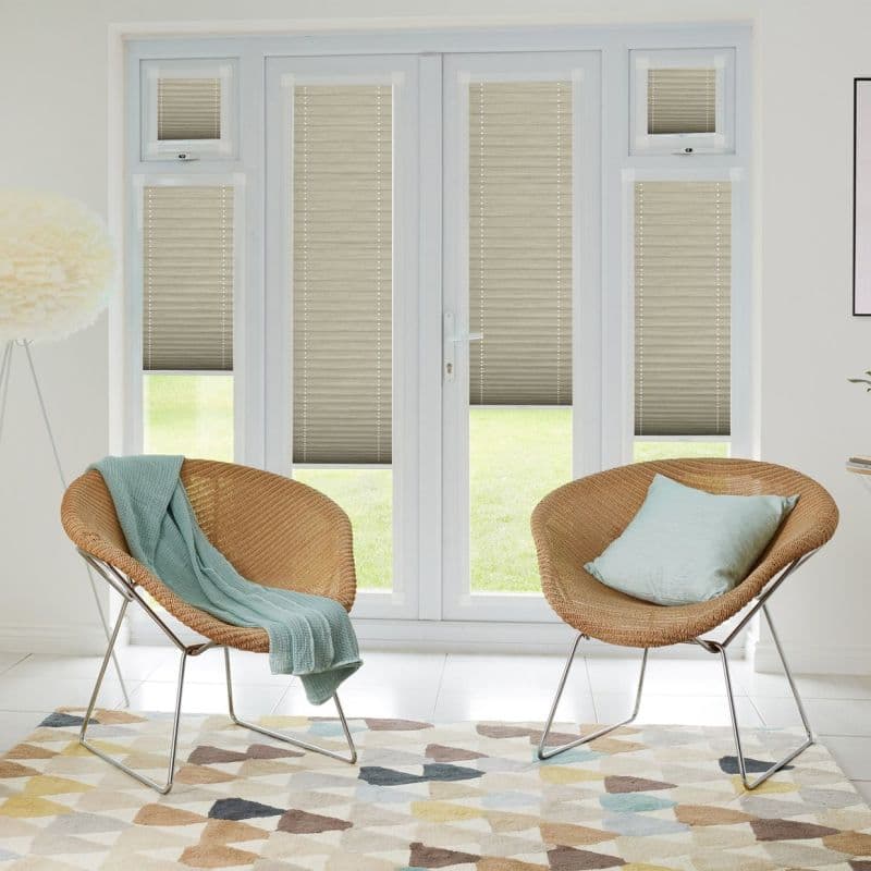 Beige pleated blinds fitted across French doors, sidelights and transoms, partially lowered to filter daylight with lower panels raised. Bright sitting room with woven chairs, cushions, patterned rug, lawn view.