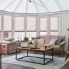 Pleated cellular blinds covering bay and roof windows, pale blush and fully closed, softly diffusing daylight; bright conservatory living area with beige sofa, coffee table, chunky pink throw and rug.