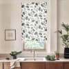 Patterned roller blind hangs over a kitchen window, blocking light with grey floral motifs; stainless-steel faucet and sink below, wooden cabinets, potted plants and a framed sketch on pale walls.
