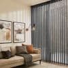Dark grey vertical blinds, floor-to-ceiling sheer slats along a corner window—filtering daylight and casting striped shadows— in a warmly lit living room with beige sofa, framed art, and wall lamp.