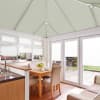 Pleated, pale-green roof blinds drawn across the pitched conservatory ceiling and white window and door blinds, filtering daylight in a bright dining-kitchen with wooden table, beige chairs and garden view.