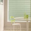 Horizontal pale-green Venetian blinds cover the window, lowered and closed, softly filtering daylight; a dining nook with a white round table, translucent lime-green chair, stool, and light wood floor.