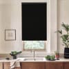 Black roller blind covers a kitchen window, blocking light; stainless faucet and sink sit beneath on a stone countertop with wooden cabinets, bowls of leafy greens and potted plants nearby.