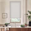 A white roller blind lowers over a kitchen window above a stainless faucet, framing a countertop with sink, bowl of greens, potted plants, a towel and wooden cabinets.