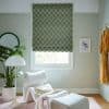 Green-and-beige checkerboard Roman blind lowered over window, fabric showing a slight bottom gap letting light through; in a pale-green sitting nook with white lounge chair, lamp, plant, framed bird print.