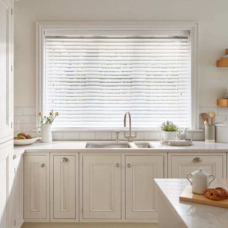White horizontal blinds (Venetian) lowered across a large window, slats closed and gently diffusing daylight; positioned above a stainless sink in a bright, neutral kitchen with cream cabinets, plants, utensils.