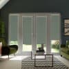 Tall glass patio doors are covered by muted-green patterned roller blinds, partially lowered, inside a living room with green armchairs, glass coffee table on a chevron rug; garden visible beneath.