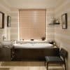 Horizontal wooden blinds centered over the window—closed, narrow slats—filter soft daylight above an oval bathtub with dark wood surround in a warm, beige-striped bathroom with sink, shelf, chair.